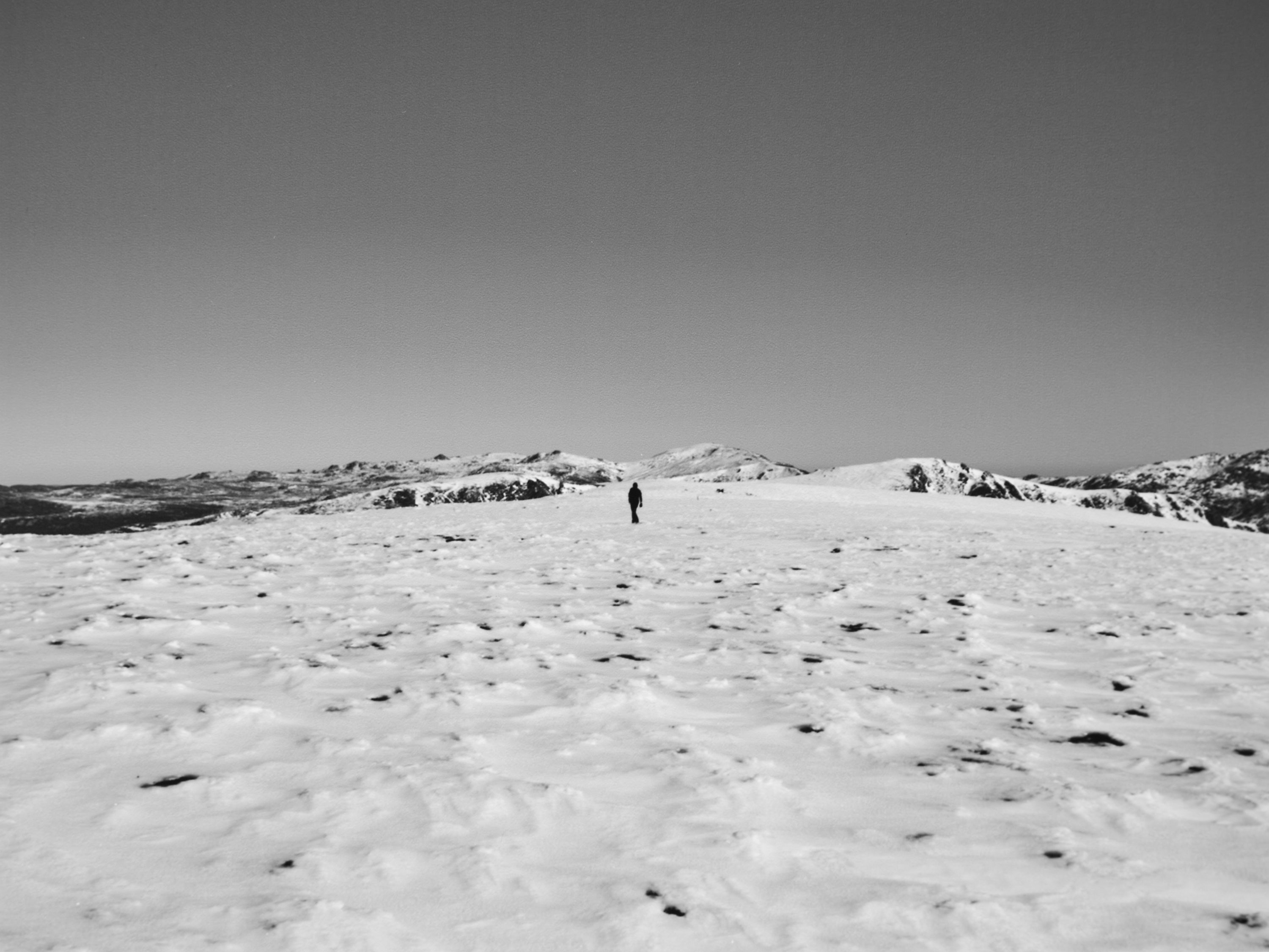 Monochrome shot of a solitary adult walking across a snow-covered mountain landscape under a clear sky, emphasizing the vastness of nature.