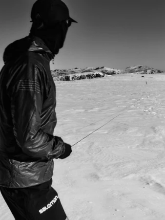 A black and white photograph of a person in sports apparel standing on a snowy field, with distant mountains visible under a clear sky.