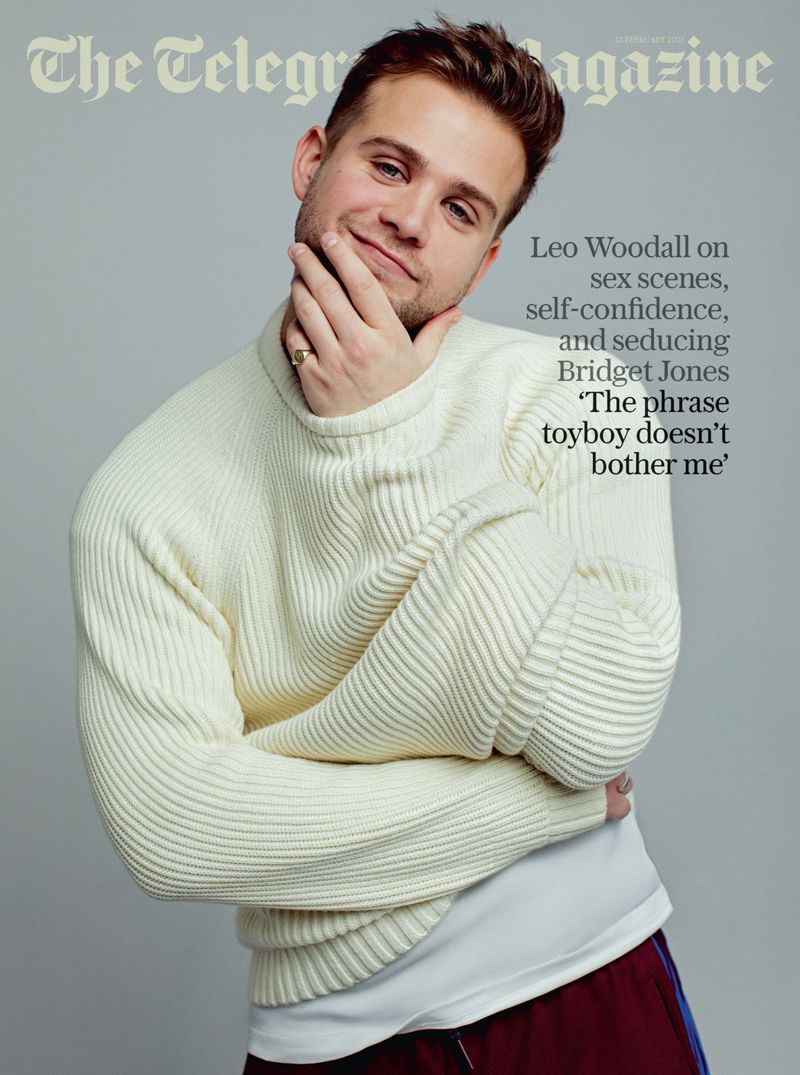 Magazine cover featuring a portrait of Leo Woodall wearing a cream sweater. He is touching his face with one hand and smiling.