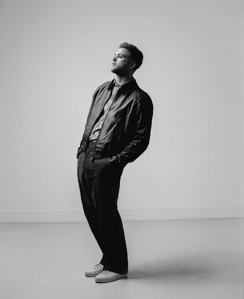 Black and white portrait of a young man in a leather jacket and dress pants leaning back slightly and looking upward in a photo studio.