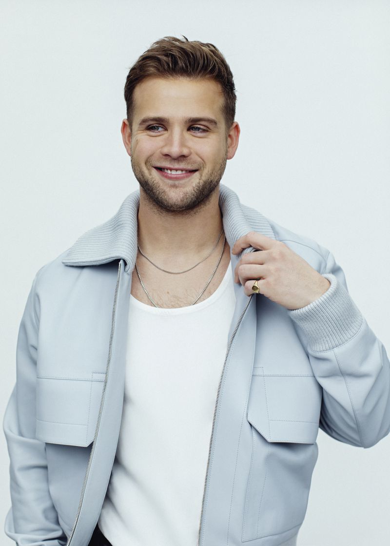 Portrait of a smiling adult male with brown hair in a light blue jacket and white t-shirt, set against a clean white backdrop.