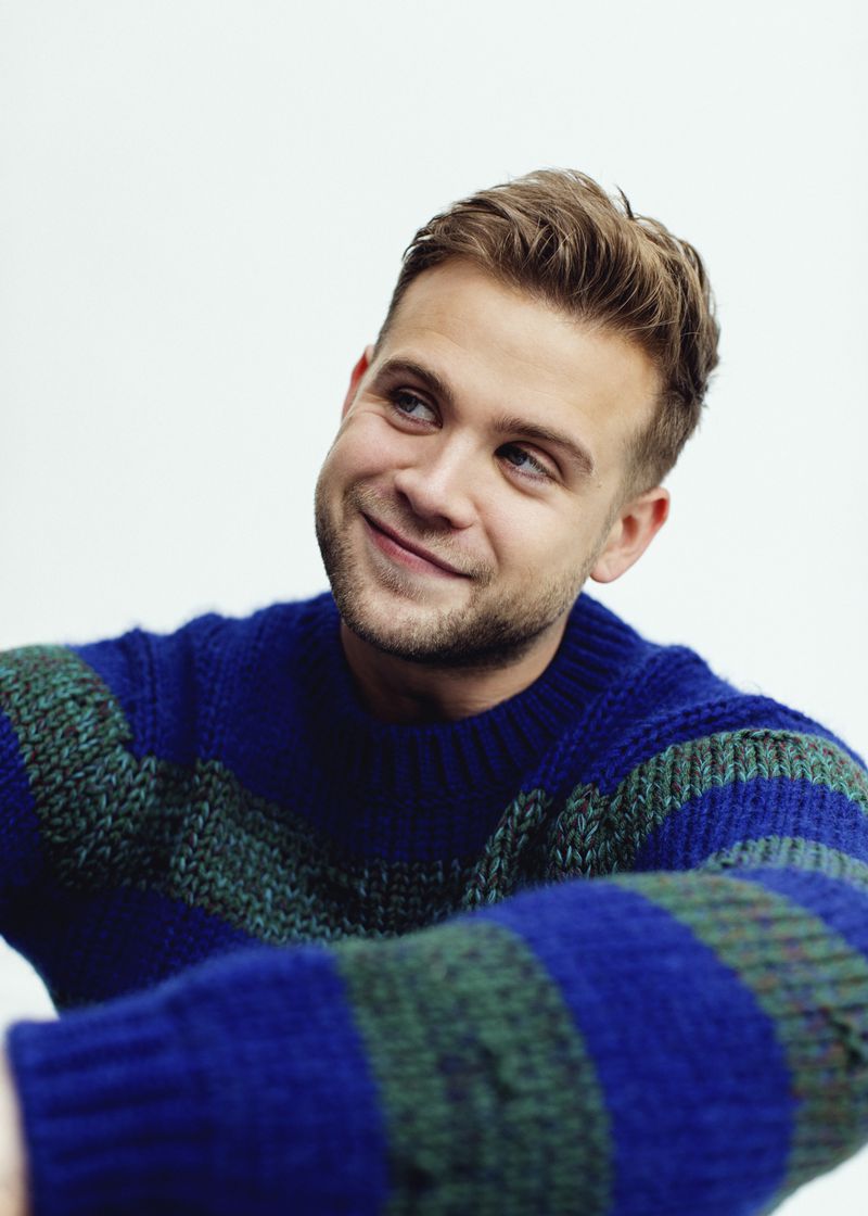 Portrait of a young adult male with light brown hair, smiling and wearing a blue and green striped sweater against a white backdrop.