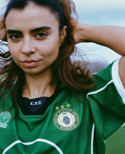 Portrait of a woman with light skin and brown hair, wearing a green and white sports jersey, outdoors under a bright sky.