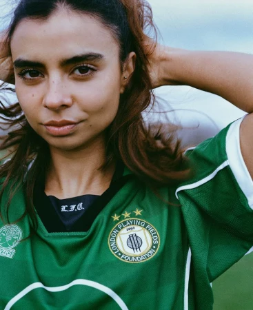 Portrait of a woman with light skin and brown hair, wearing a green and white sports jersey, outdoors under a bright sky.