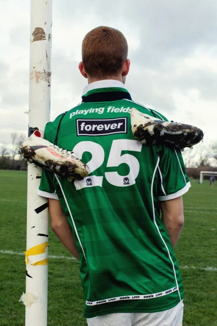 Back view of a person in a green sports jersey with shoes draped over the shoulders, leaning against a white pole on a field.