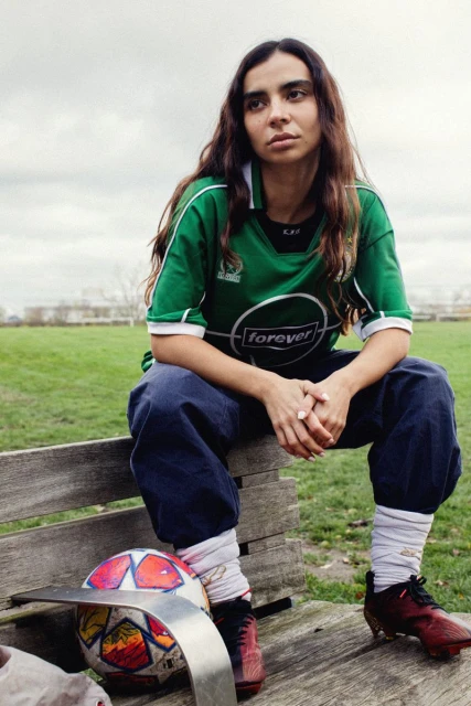 Portrait of a young adult woman in a green soccer jersey sitting on a park bench with a soccer ball in an outdoor setting.