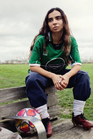 Portrait of a young adult woman in a green soccer jersey sitting on a park bench with a soccer ball in an outdoor setting.