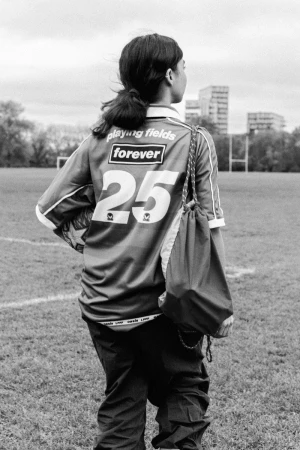Teenager stands on a field, back to the camera, wearing soccer jersey, bag on her shoulder, looking at buildings in distance.