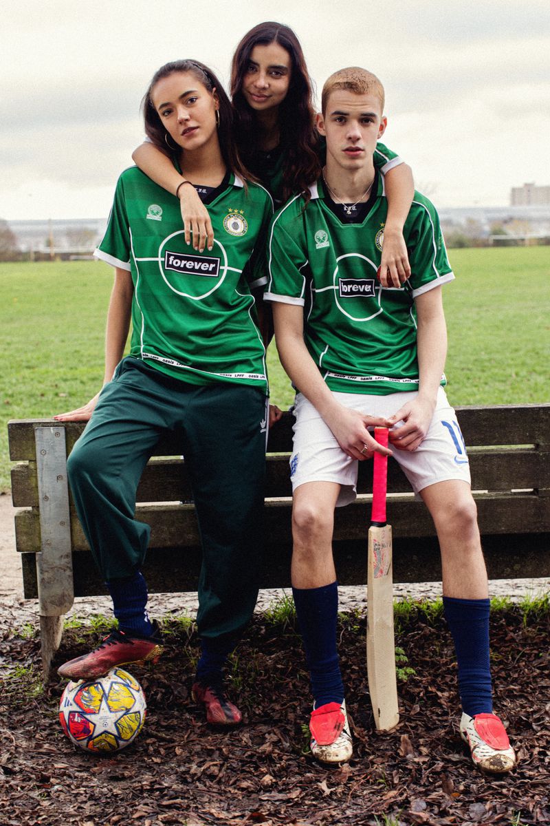 Three teenagers in matching green sports jerseys pose together in an outdoor park. One holds a cricket bat and soccer ball.