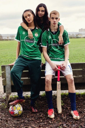 Three teenagers in matching green sports jerseys pose together in an outdoor park. One holds a cricket bat and soccer ball.