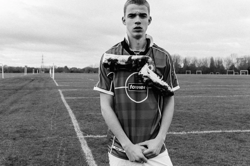 Black and white portrait of a young adult male in a soccer jersey with soccer cleats around his neck on a grass field.