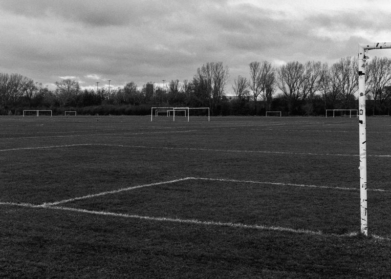 Monochrome shot of a soccer field with grass and white line markings under a cloudy sky, multiple soccer goals visible in the background.