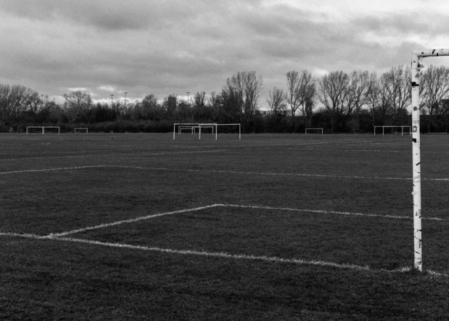 Monochrome shot of a soccer field with grass and white line markings under a cloudy sky, multiple soccer goals visible in the background.