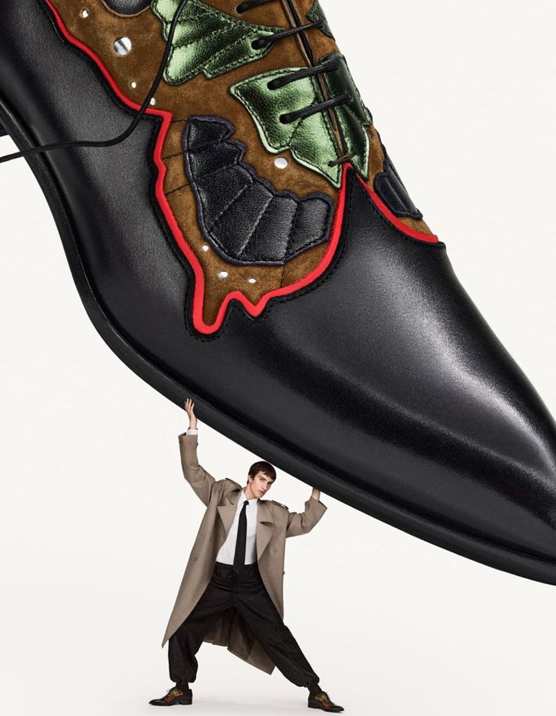 A man in a tan trench coat struggles humorously under the weight of an oversized black shoe with brown and green decorative patches.