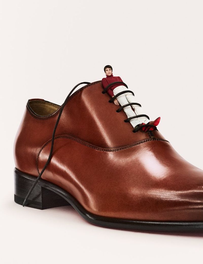 Conceptual fashion shot of a small male figure posed on the laces of a large brown leather dress shoe against a plain white background.