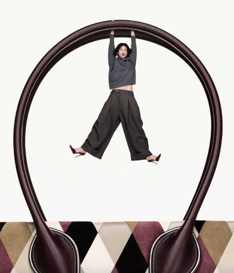 Fashion shot featuring a woman in a grey sweatshirt and wide-legged pants hanging from the handle of a large handbag against a white backdrop.