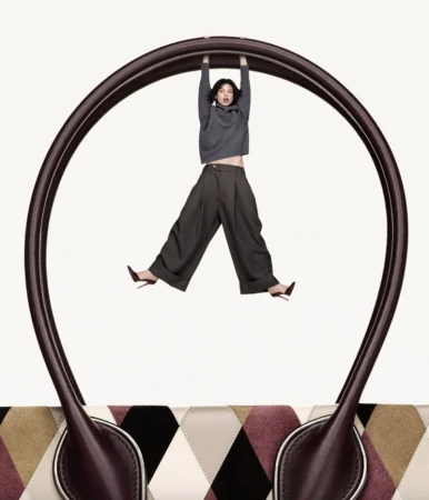 Fashion shot featuring a woman in a grey sweatshirt and wide-legged pants hanging from the handle of a large handbag against a white backdrop.