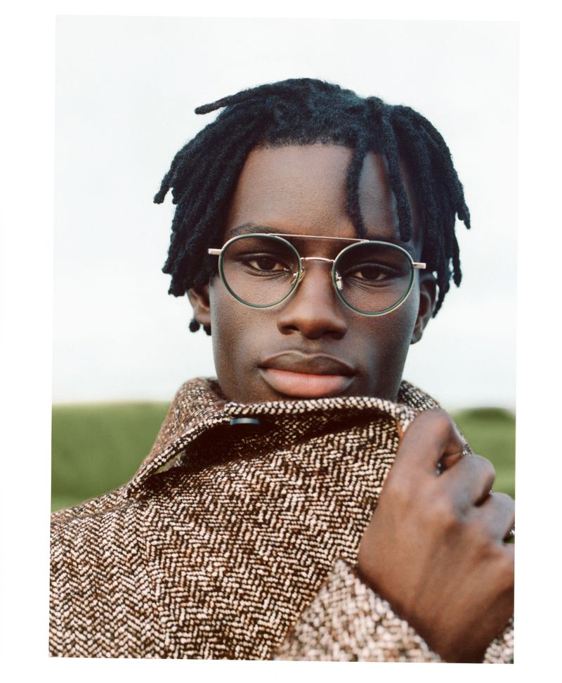 Close-up portrait of a Black adult male wearing glasses and a patterned tweed jacket, set against a blurred outdoor backdrop.