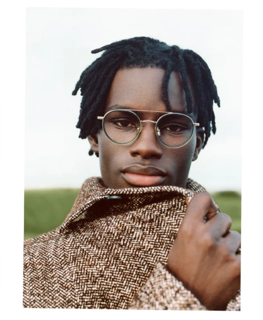 Close-up portrait of a Black adult male wearing glasses and a patterned tweed jacket, set against a blurred outdoor backdrop.