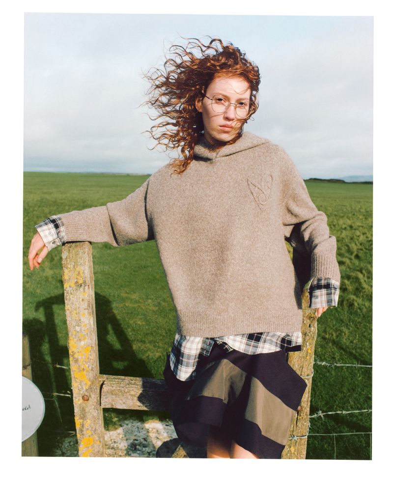 Red-haired woman in a beige knit hoodie leaning against a weathered wooden post in an open grassy field under an overcast sky.