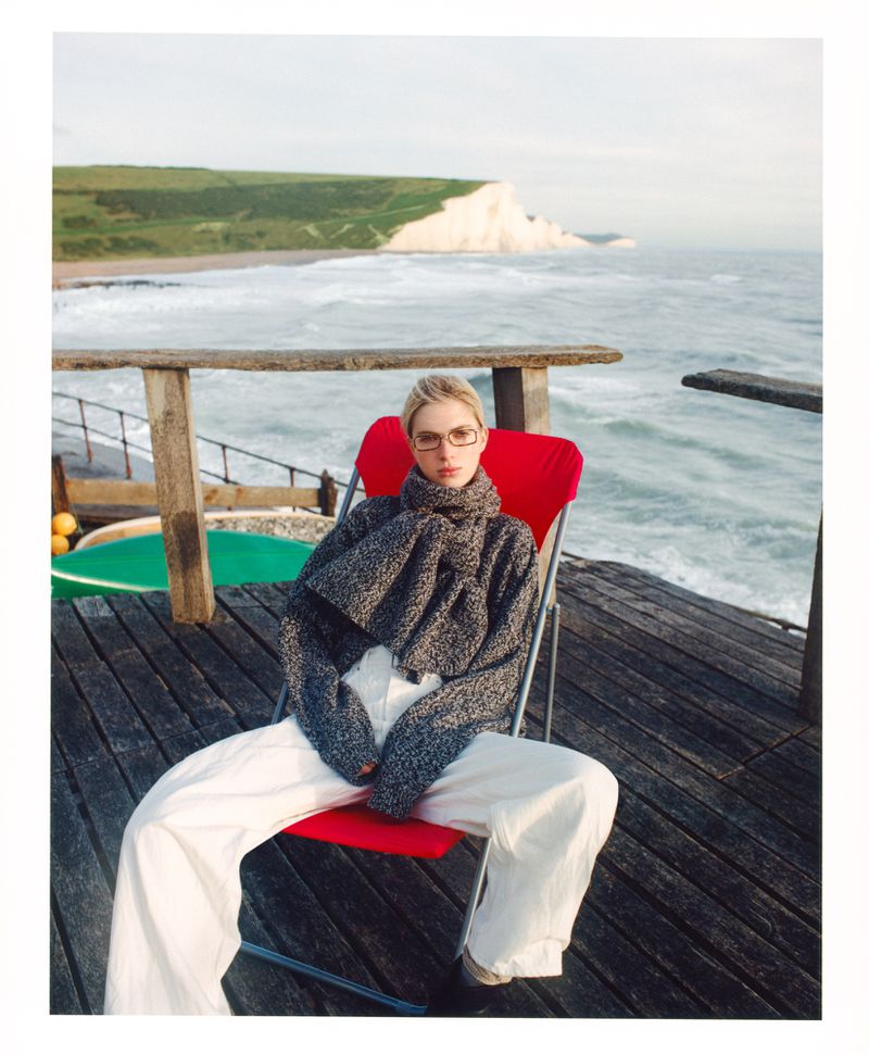 A woman sits on a red chair on a wooden deck overlooking the ocean with white cliffs in the distance, wearing a grey sweater and white pants.
