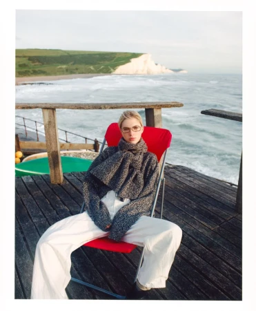 A woman sits on a red chair on a wooden deck overlooking the ocean with white cliffs in the distance, wearing a grey sweater and white pants.