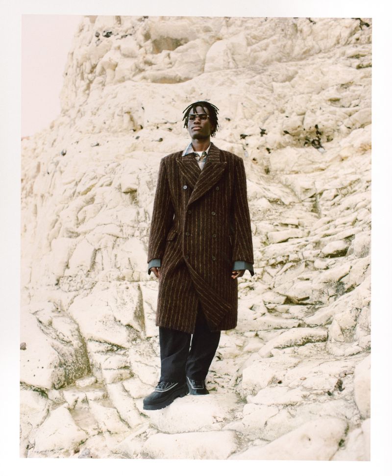 Man wearing a brown pinstripe double-breasted coat standing against a textured rocky white cliff background