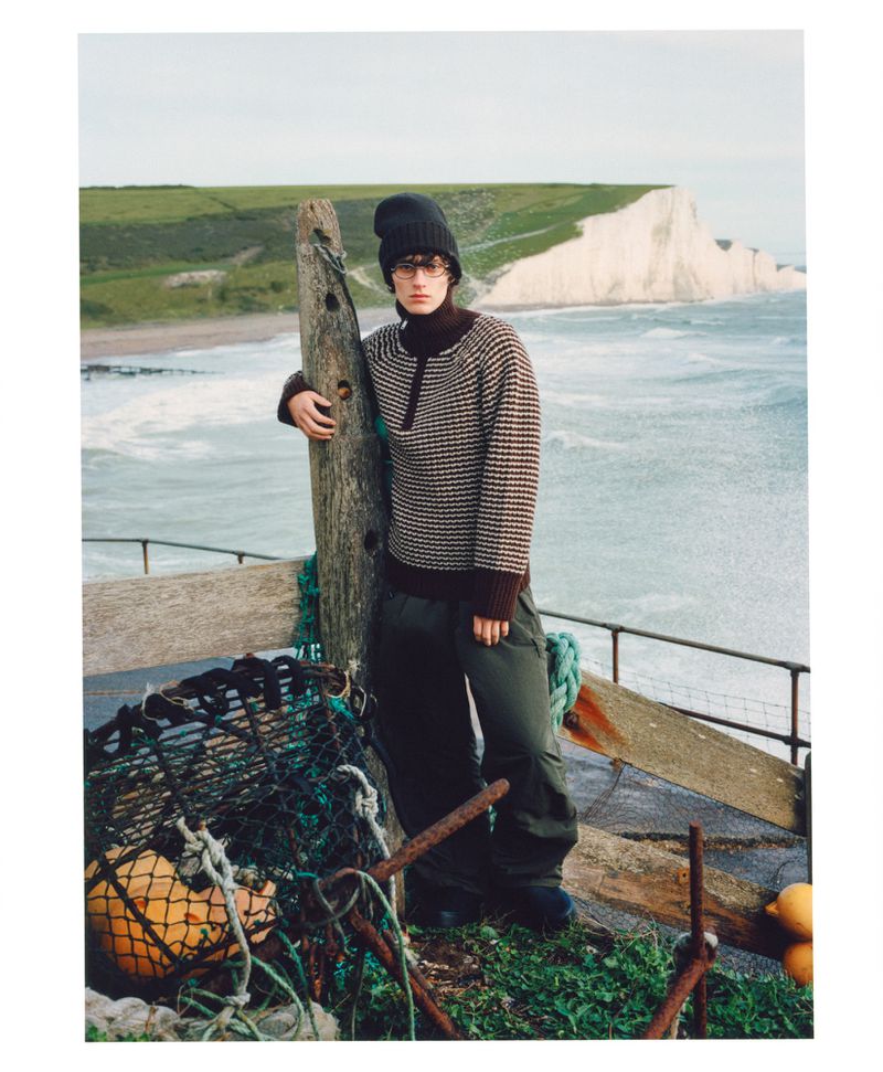 A young adult stands on a weathered wooden structure against a backdrop of the ocean and chalk cliffs, wearing a patterned sweater.