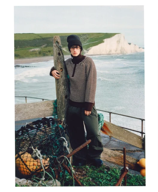 A young adult stands on a weathered wooden structure against a backdrop of the ocean and chalk cliffs, wearing a patterned sweater.