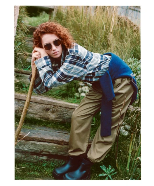 A woman with auburn curly hair, wearing a plaid shirt, leans on a wooden railing in a grassy, outdoor setting while sporting sunglasses.