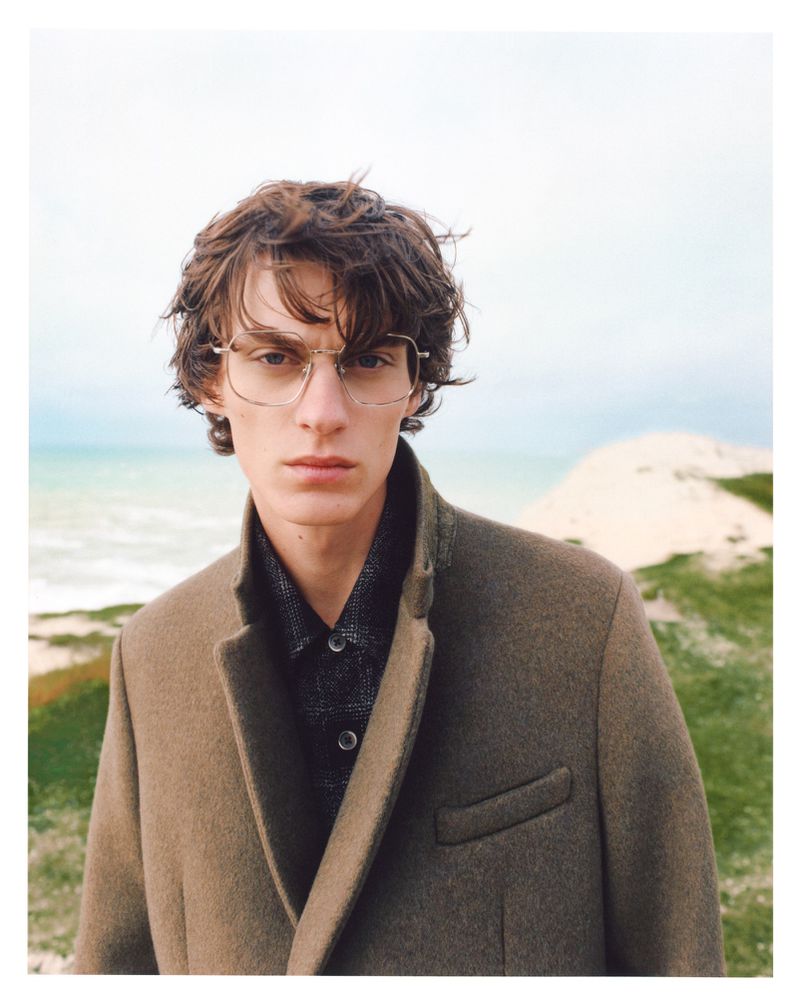 Adult male with eyeglasses and brown coat posing outdoors against a diffused, bright background of dunes, beach, and sky.