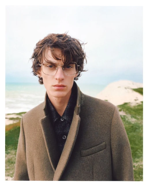 Adult male with eyeglasses and brown coat posing outdoors against a diffused, bright background of dunes, beach, and sky.