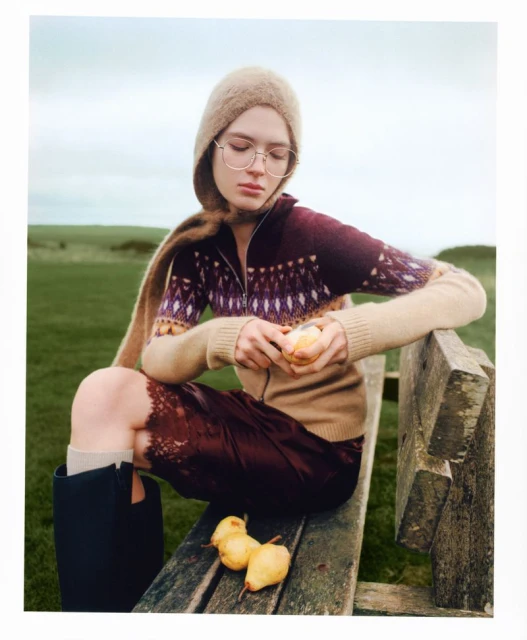 Woman in wool hat and sweater sits on a wood bench outdoors, peeling fruit with three pears next to her, soft diffused light.