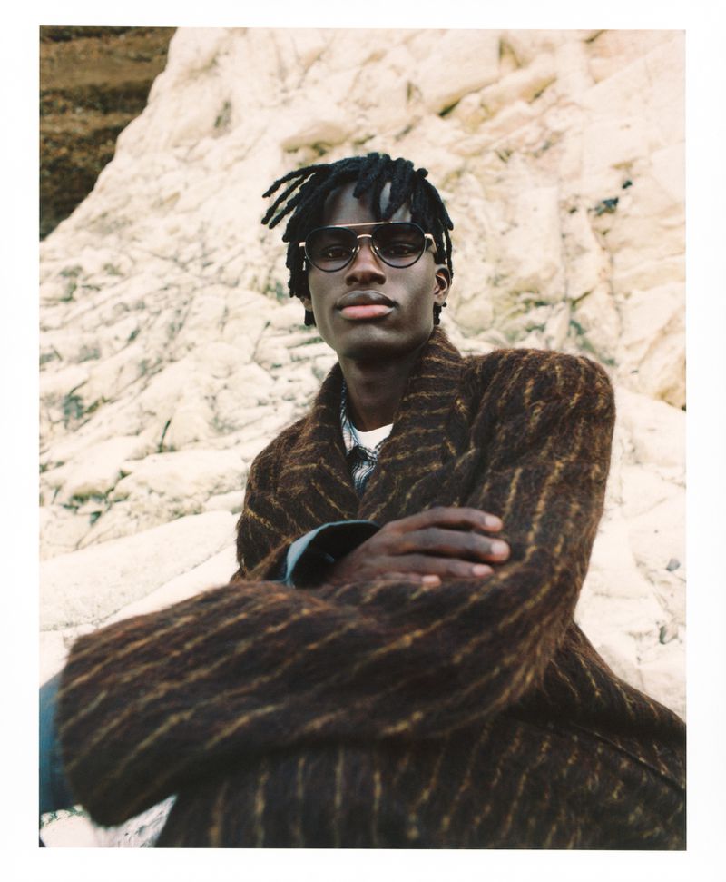 A dark-skinned man with dreadlocks, wearing sunglasses and a brown-striped coat, poses outdoors with a textured rock formation as a backdrop.