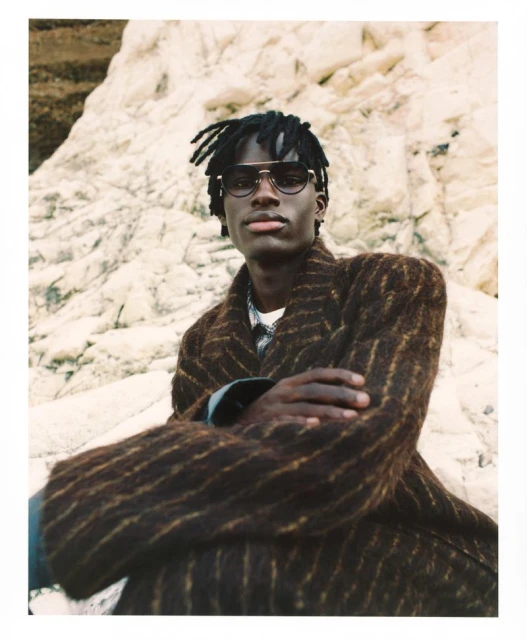A dark-skinned man with dreadlocks, wearing sunglasses and a brown-striped coat, poses outdoors with a textured rock formation as a backdrop.