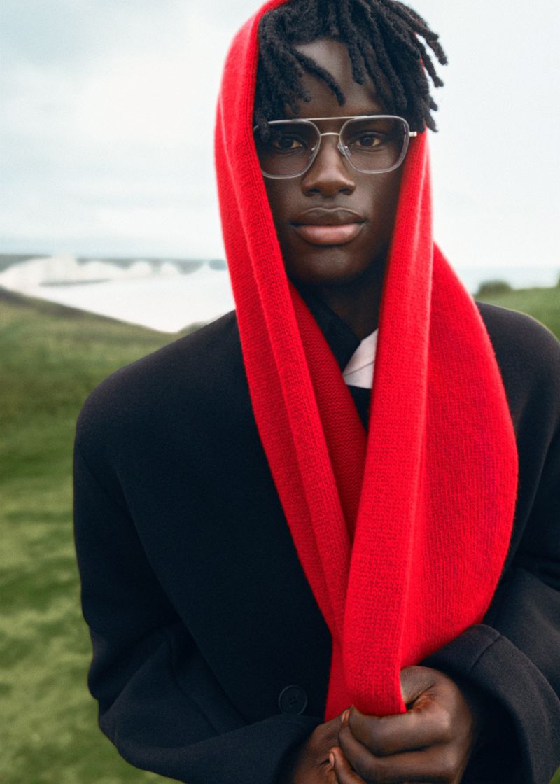 Close-up of a stylish Black man wearing a red scarf and glasses outdoors, with a blurry green and white background.