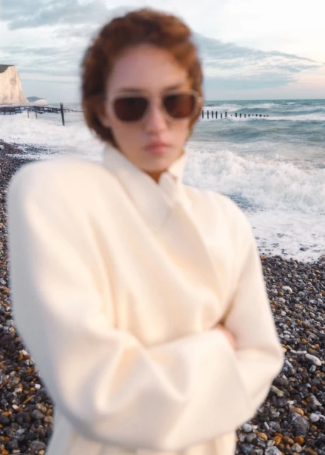 Adult woman wearing sunglasses and a cream-colored coat standing on a rocky beach with ocean waves and a chalk cliff background.