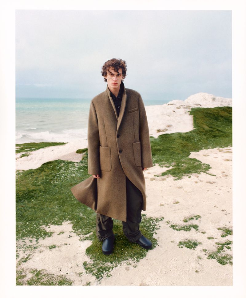 A young adult man stands in a brown wool overcoat and black boots on a grassy hill overlooking the ocean on an overcast day.