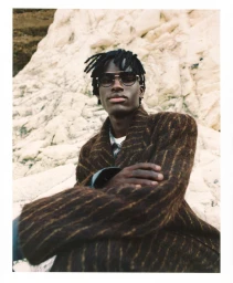 Black man wearing dark sunglasses and a textured brown coat seated against a rocky white background