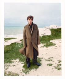Man wearing a long brown coat standing on a grassy coastal shore near the sea for Bloobloom Winter 2025 by Luc Coiffait