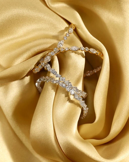 Close-up of two diamond bracelets, one gold, one silver, intertwined on a draped gold silk fabric background, showcasing luxury jewelry.