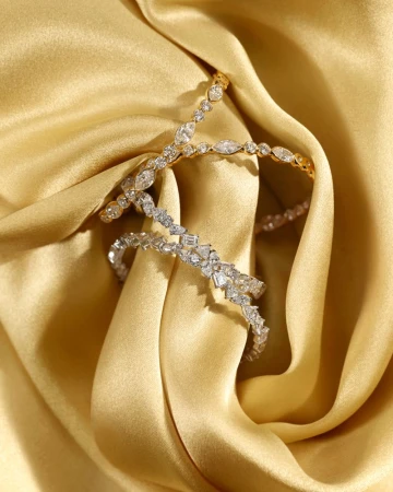 Close-up of two diamond bracelets, one gold, one silver, intertwined on a draped gold silk fabric background, showcasing luxury jewelry.