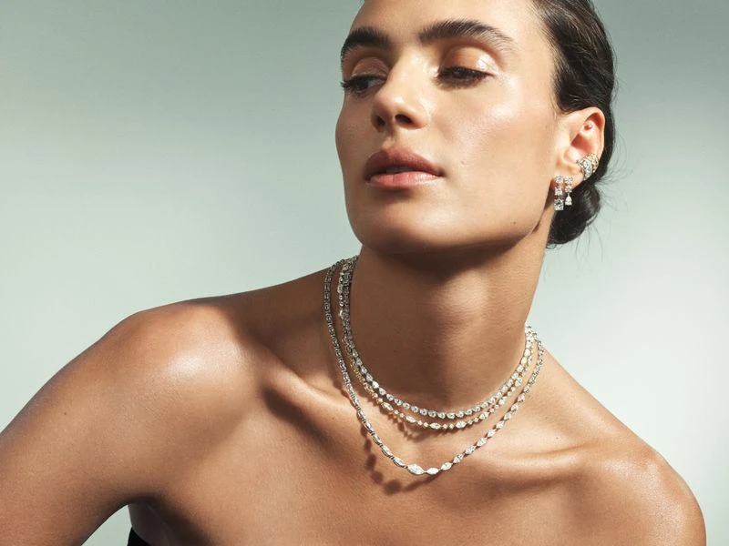 Close-up of a woman with her shoulders bare, wearing layered necklaces and earrings against a light green background in a studio.