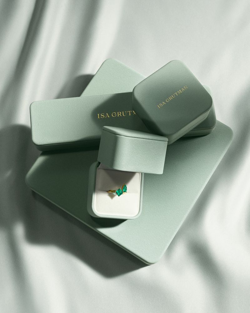 Elegant presentation of a gold ring with green gems, nestled in a light green jewelry box against a soft, white, draped fabric backdrop.