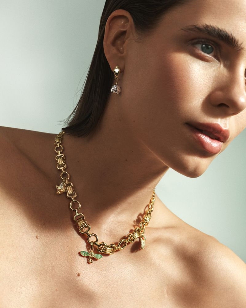 Close-up of a woman wearing a gold statement chain necklace with charms and drop earrings, shot indoors under soft, diffused lighting.