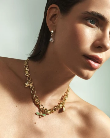 Close-up of a woman wearing a gold statement chain necklace with charms and drop earrings, shot indoors under soft, diffused lighting.