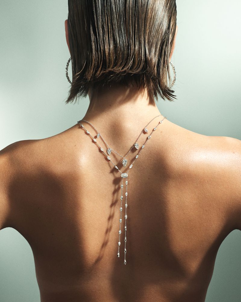A back shot of an adult woman with short brown hair, wearing a diamond necklace and hoop earrings, against a light teal background.