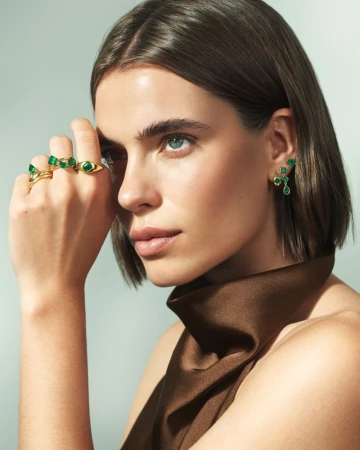 Close-up of a woman wearing green gemstone rings and earrings, with brown hair and a brown dress, in a studio setting.