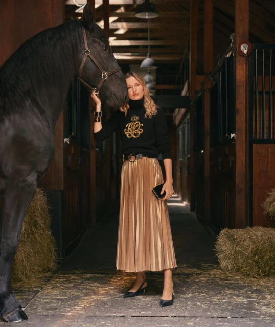 A woman in a black sweater and gold pleated skirt stands in a horse stable next to a large black horse. Fashion and equestrian.