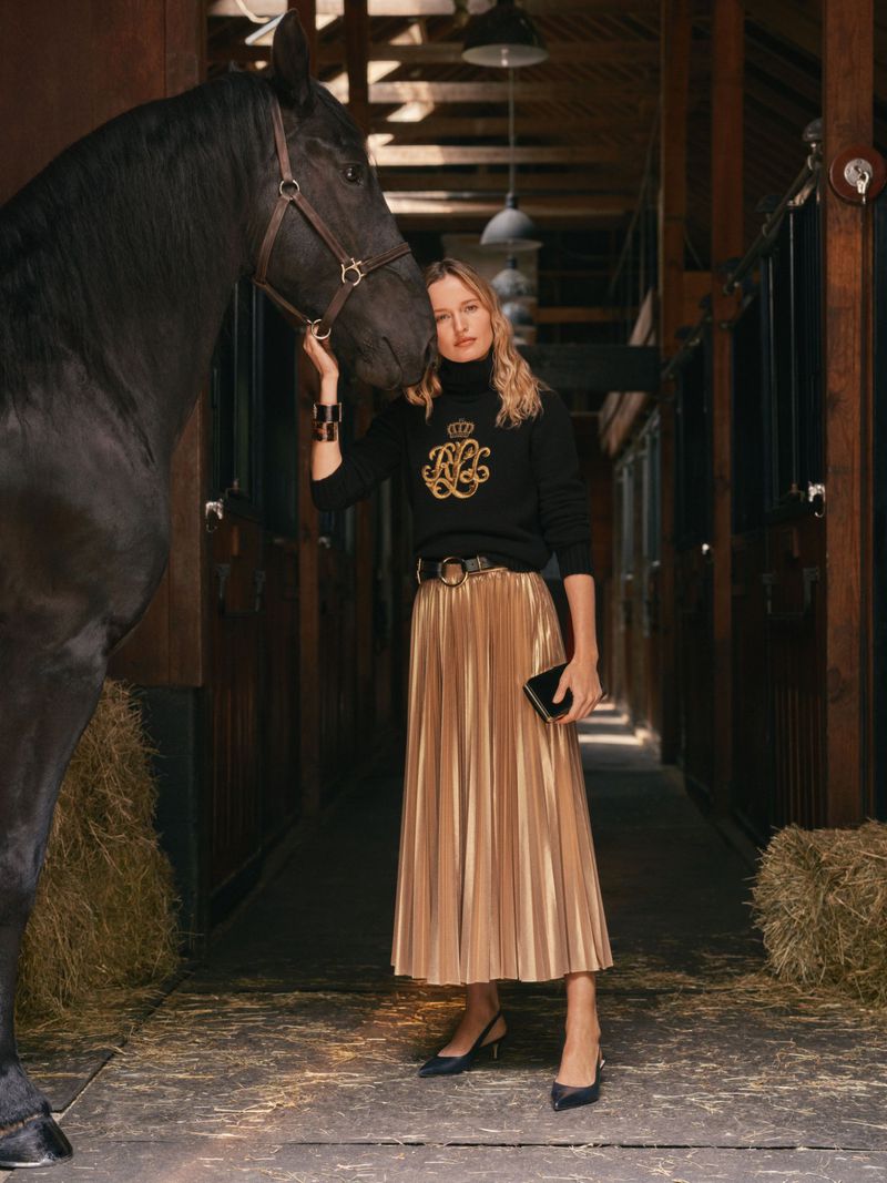 Woman posing with a black horse inside a stable. The model wears a black sweater, pleated gold skirt, and heels in a fashion shot.